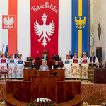 widok prezydium na sali Sejmu wokoło Przewodniczacęgo i Wiceprzewodniczących stoją artyści w strojach ludowych