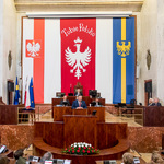 widok na prezydium sali Sejmu z oddali, mężczyzna przemawia na mównicy w bocznych ławach zasiadają goście