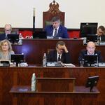 mężczyzna siedzący za biurkiem przemawia, poniżej siedzą trzy osoby