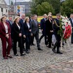 Grupa ludzi kroczy w stronę pomnika przed nimi żołnierz niesie wieniec.